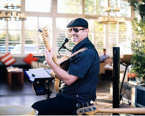 A musician, wearing a cap and sunglasses, smiles while playing an electric guitar with a microphone in front of him at what seems to be an outdoor venue.