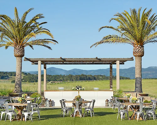 The image shows an outdoor event setup with tables and chairs under palm trees and a pergola, set in a scenic landscape with mountains in the background.