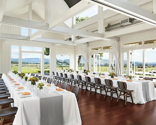 A bright, elegant dining room with large windows, set for an event with long tables, white tablecloths, gray runners, and neatly arranged chairs.