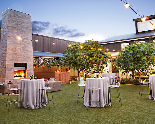 An outdoor event space with tall cocktail tables, a fireplace, string lights, trees, and a modern building in the background, during twilight.