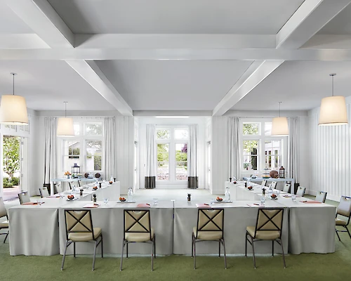 A bright, modern conference room setup with a U-shaped table arrangement, white tablecloths, chairs, hanging lights, and large windows.