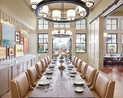 A long dining table with beige chairs, set with plates and wine glasses, situated under a large chandelier in a well-lit, elegant dining room.