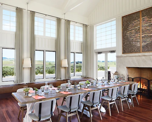 A bright dining room with a long table, multiple chairs, large windows, a fireplace, and modern decor overlooking a scenic outdoor view.