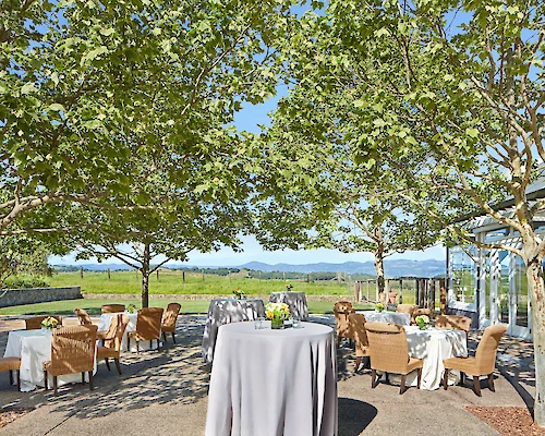 An outdoor dining area is set up with tables and chairs under trees, offering a scenic view of the mountains in the distance.