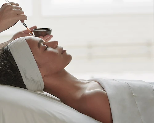 A person is laying on a spa bed with a headband, receiving a facial treatment while a practitioner applies a mask with a brush.