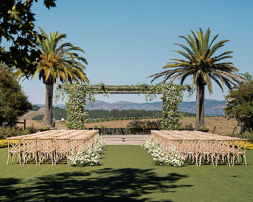 An outdoor wedding setup features rows of wooden chairs facing a green arch adorned with foliage. Palm trees, mountains, and clear skies surround the scene.