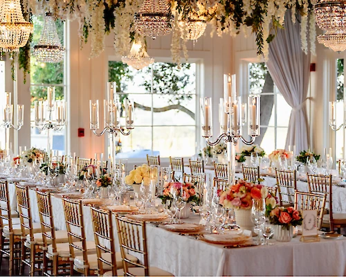 The image shows an elegantly decorated wedding reception with long tables, floral centerpieces, chandeliers, and candelabras in a bright, airy venue.