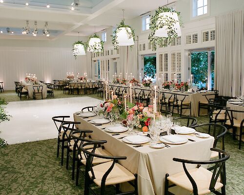 An elegantly decorated dining hall with arranged tables, floral centerpieces, and hanging lights with greenery, ready for an event.