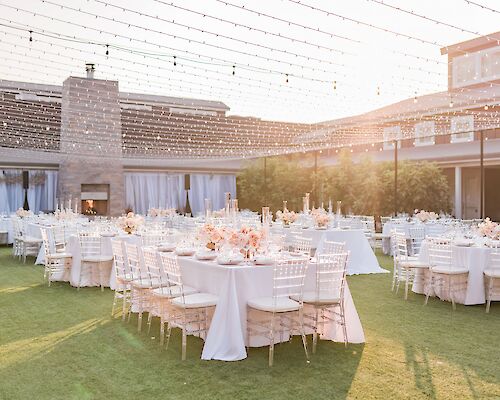 An elegant outdoor event setup features round tables with white tablecloths and decor, surrounded by white chairs, under string lights.