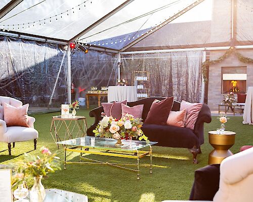 A cozy, elegant tent with white chairs, a black sofa, a glass coffee table, and floral arrangements on a grassy floor.