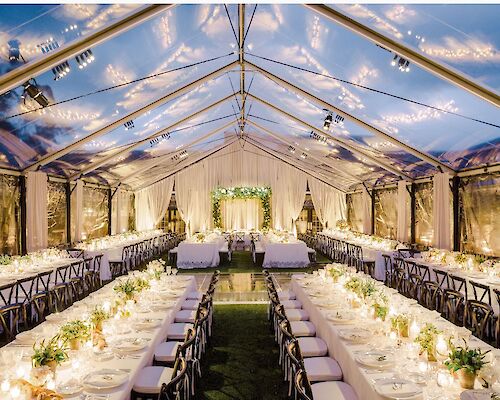 The image shows a beautifully decorated wedding reception under a transparent tent with long dining tables, floral arrangements, and string lights.