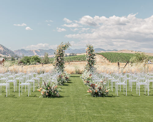 An outdoor wedding setup with clear chairs and a floral arch on green grass, surrounded by scenic hills and vineyards under a partly cloudy sky.