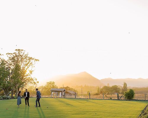 Three people stand on a large, grassy field at sunset with trees and a small building in the background, and mountains in the distance.