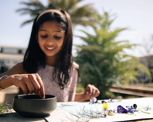 A girl is seated outdoors, engaging in an activity with a bowl and surrounding flowers and greenery on a table.