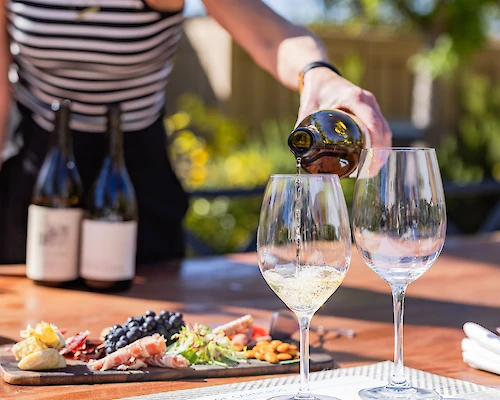 A person pouring wine into glasses on a table set with charcuterie and other food items, with wine bottles in the background.